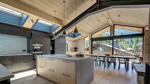 A modern kitchen with a central island, wooden ceiling, and large windows offering views of a wooden deck and trees. - Chic & Spacious Chalet with Hot Tub & Sauna (Chamonix-Mont-Blanc)