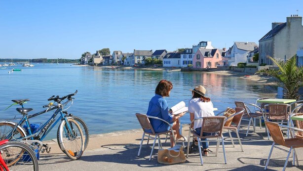 A waterfront café with outdoor seating, a bicycle, and a view of the water and buildings. - 🌊 Ti Glaz - Île-Tudy – A charming home between the sea and the river (Île-Tudy)