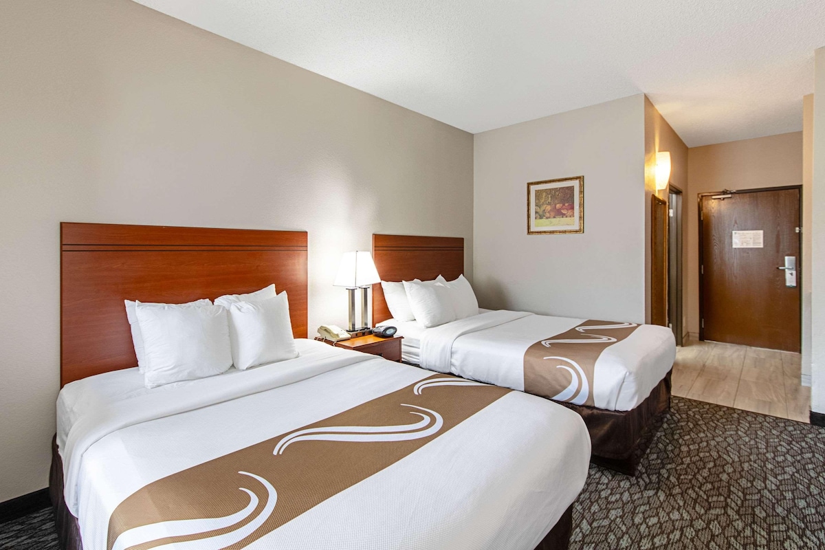 A hotel room with two beds, wooden headboards, white bedding, a lamp, a telephone, and a framed picture on the wall.