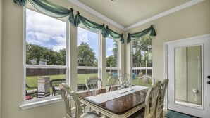 Zona pranzo con tavolo in legno, sedie bianche e vista sul giardino retrostante attraverso ampie finestre.