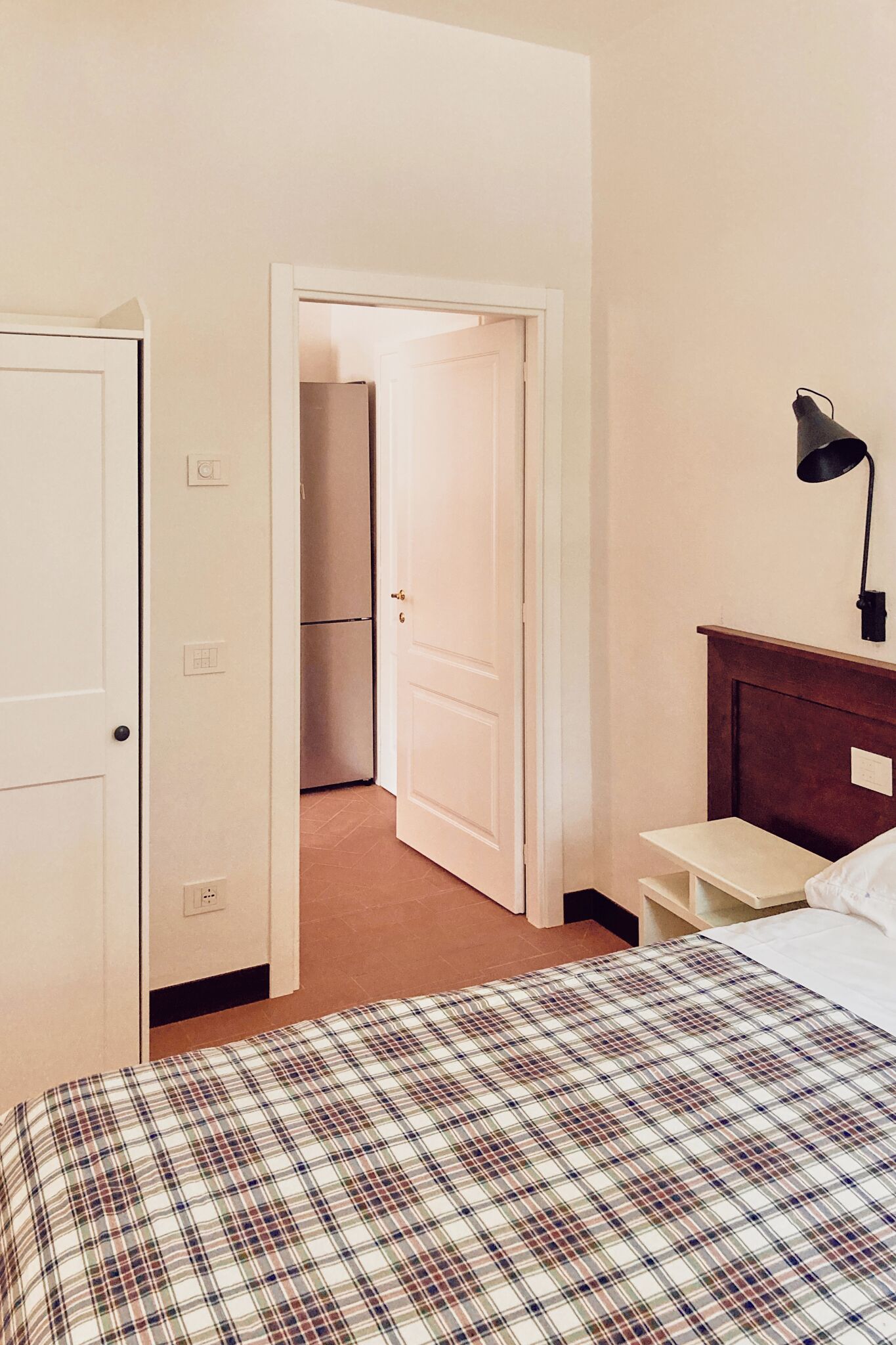 Une chambre avec une couvre-lit à carreaux, une table de chevet en bois, une lampe noire et une porte blanche donnant sur un couloir.