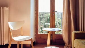 Una habitación con una silla de madera, una mesita y un sofá. Se ve una ventana con cortinas y vistas al exterior con vegetación.