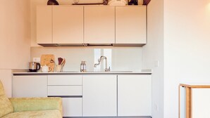 Una cocina compacta con armarios blancos, una encimera y un lavamanos. En la encimera hay una tabla de cortar de madera, un hervidor y una cafetera.