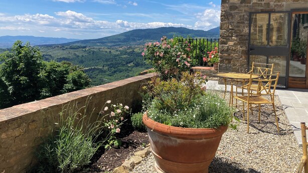 Una terraza con un muro de piedra, una maceta de terracota grande con plantas, una mesa de mimbre con sillas, y vistas a un paisaje montañoso.