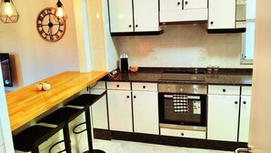 A modern kitchen with white cabinets, a wooden countertop, and a built-in oven. There are hanging pendant lights and a clock on the wall. - La Terraza de Alma (O Milladoiro)