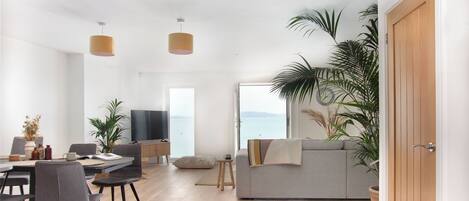 Amplia sala de estar con mesa de comedor, sillas, sofá y televisor. Hay plantas en macetas y vistas al mar a través de amplias ventanas.
