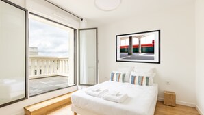 Une chambre moderne avec une grande fenêtre, un lit sur lequel sont soigneusement pliées des serviettes, et un tableau encadré accroché au mur.