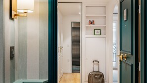 Un couloir d’hôtel avec un plancher en bois, une valise, une lampe fixée au mur et une porte dotée d’un cadre vert.