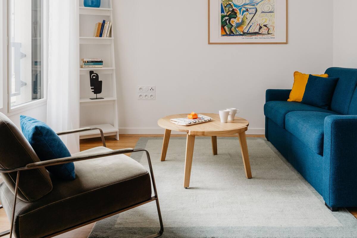 Un salon moderne avec un canapé bleu, une table basse en bois et un fauteuil noir. On y trouve une bibliothèque remplie de livres et une œuvre d’art encadrée accrochée au mur.