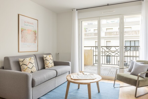 A modern living room with a grey sofa, a round wooden table, and a balcony with a view of a building. - Residence Edgar Suites Levallois - Jules Verne (Levallois-Perret)