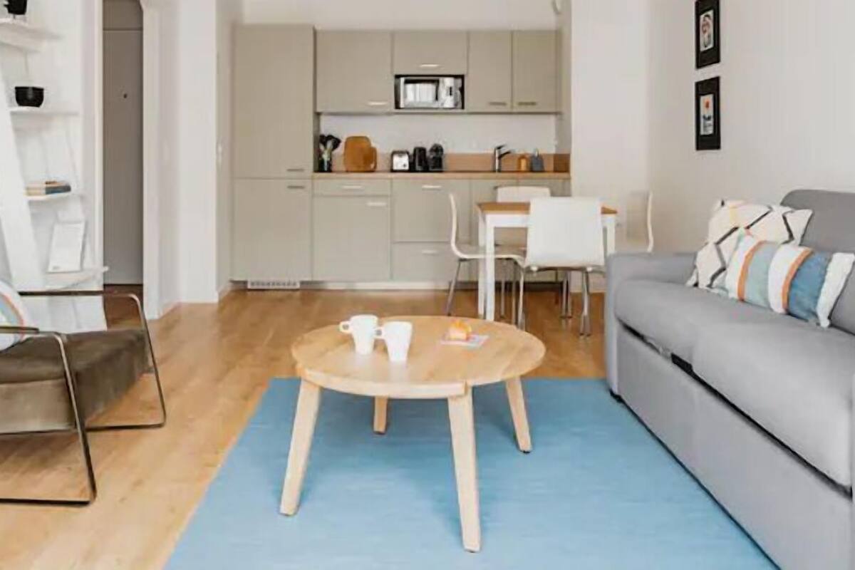 Un salon moderne avec un canapé gris, un fauteuil marron et une table basse en bois. On aperçoit à l’arrière-plan une cuisine avec des meubles blancs et un coin repas comprenant une table et des chaises blanches.