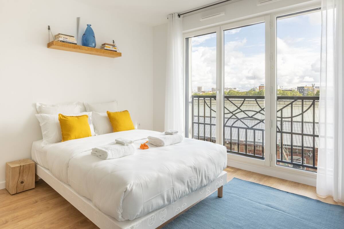 Une chambre avec un grand lit, une table de chevet en bois, une étagère avec des livres et un vase, et un balcon avec une balustrade.