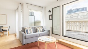 Un salon moderne avec un canapé gris, une petite table en bois et une grande fenêtre donnant sur un immeuble.