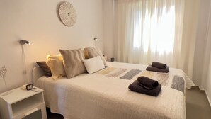 Una cama bien hecha con edredón blanco y varios almohadones, una mesita de noche con un reloj y un teléfono, y un reloj de pared.