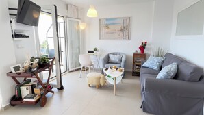 Una sala moderna con un sofá gris, una mesita redonda pequeña y una silla blanca. Hay un televisor montado en la pared, un cuadro sobre el sofá y una puerta corrediza de vidrio que da a un espacio exterior.