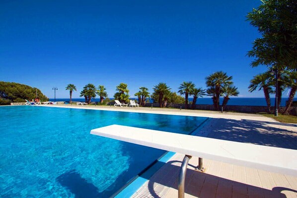 A clear blue swimming pool with a white edge, surrounded by palm trees and lounge chairs, with a view of the sea. - Casa delle Margherite 1 (Piani di Cipressa)