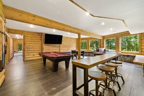 A wooden cabin interior with a pool table, bar stools, and a TV mounted on the wall.