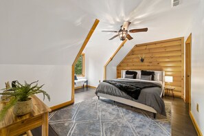 A bedroom with a sloped ceiling, a bed with a dark gray blanket, a wooden headboard, a small table with a plant, and a nightstand with a lamp.