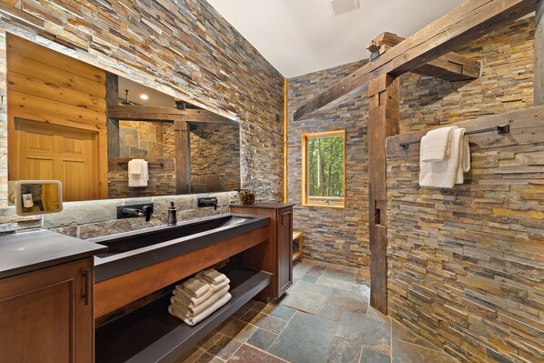 A bathroom with a stone wall, wooden beams, a large mirror, a double sink, and a towel rack.