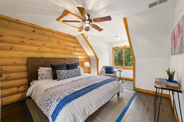 Une chambre avec un plafond en bois, un lit avec une literie à motifs, un ventilateur de plafond, une fenêtre, une chaise et une petite table sur laquelle est posée une plante.