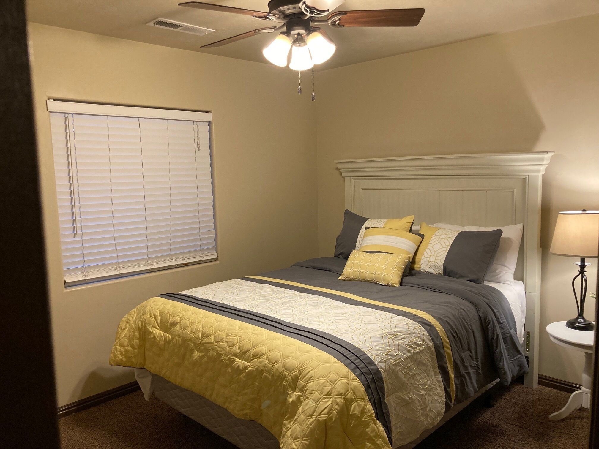 Une chambre à coucher comprenant un lit, un ventilateur de plafond, une fenêtre avec des stores et une table de chevet avec une lampe.