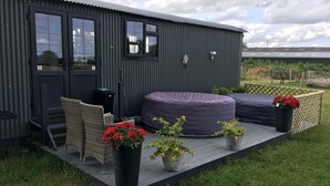 A compact, dark-colored building with a deck, outdoor seating, and potted plants. - Charming Luxurious Rural Shepherds Hut (Hook)