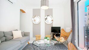 Uma sala de estar moderna com um sofá cinzento, uma mesa de centro de vidro e uma televisão. Há dois espelhos hexagonais na parede, uma poltrona de vime e uma janela com cortinas.