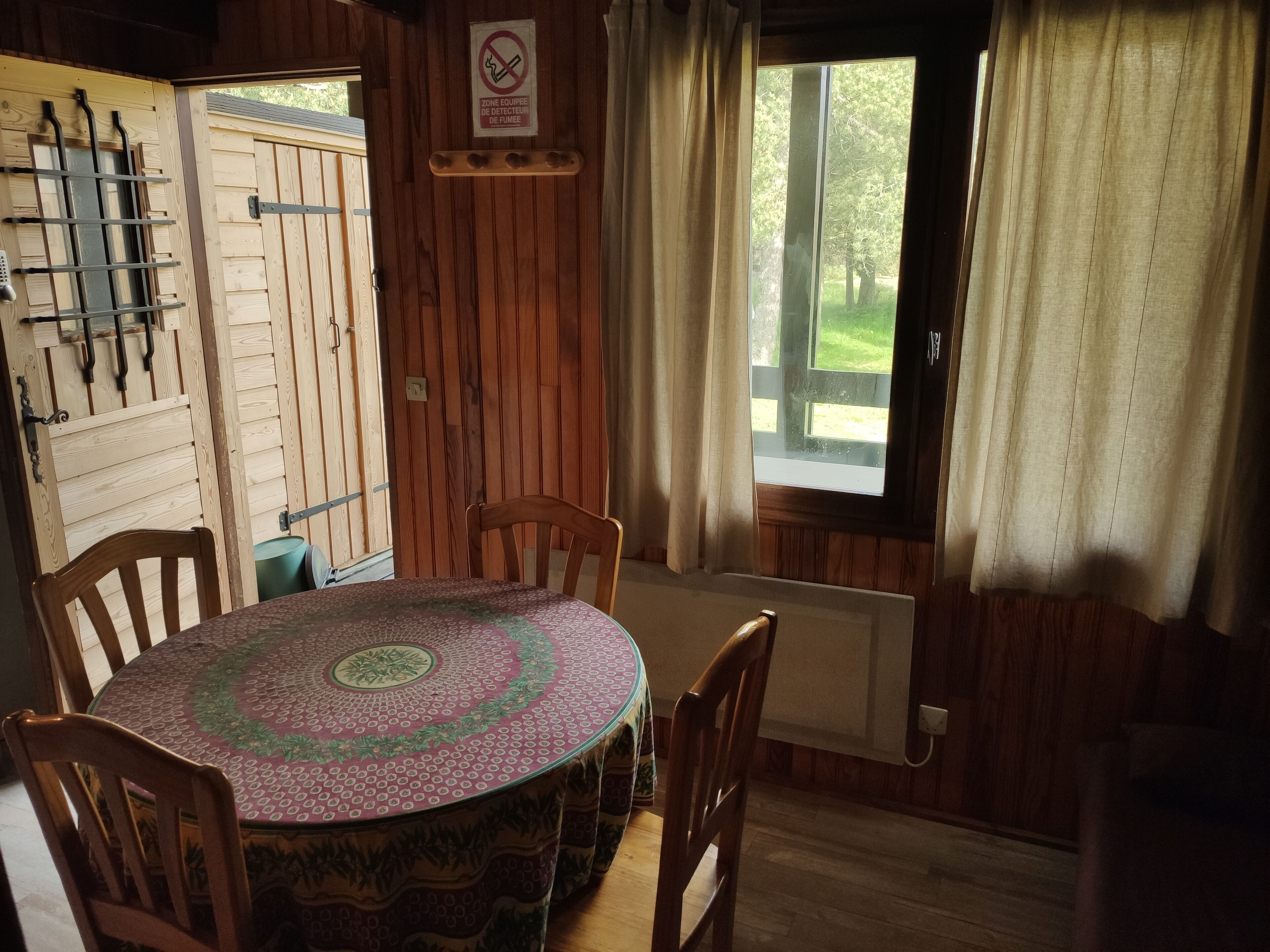A wooden interior with a round table, chairs, a window with curtains, and a sign indicating a smoking area.