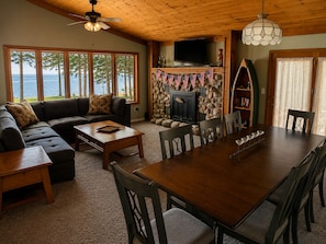 A cozy living room with a fireplace, a stone wall, a large window overlooking the sea, a sofa, and a dining table with chairs. - Waterfront on Lake Huron- NEW Kitchen & Bathroom (Cheboygan)