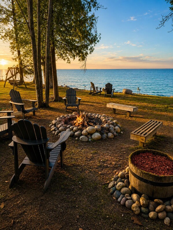 A lakeside outdoor seating area with Adirondack chairs, a fire pit, and a wooden bench.