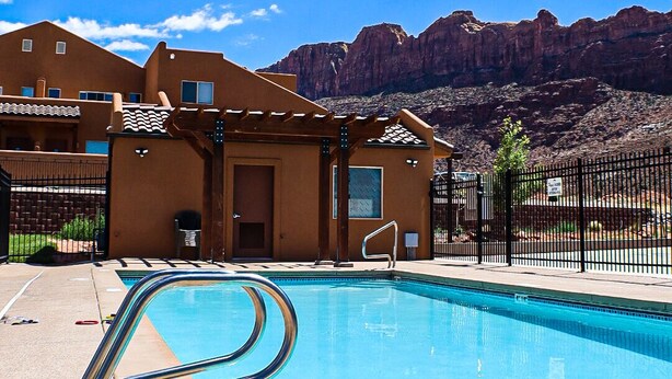 A swimming pool with a metal ladder, a covered patio area, and a mountain backdrop. - Desert Willow Rim Vista 6A7 (Moab)
