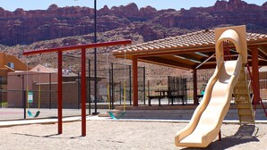 A playground with a slide, swings, and a covered seating area, set against a mountainous backdrop. - Desert Willow Rim Vista 6A7 (Moab)