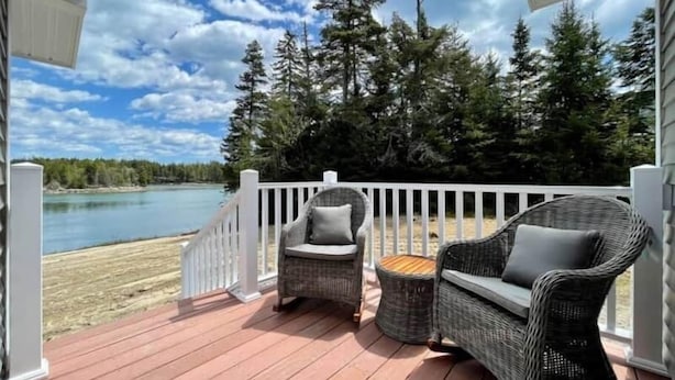 A wooden deck with wicker furniture, a white railing, and a view of a lake and forest. - Waterfront Cottage on Private Island near Acadia (Milbridge)