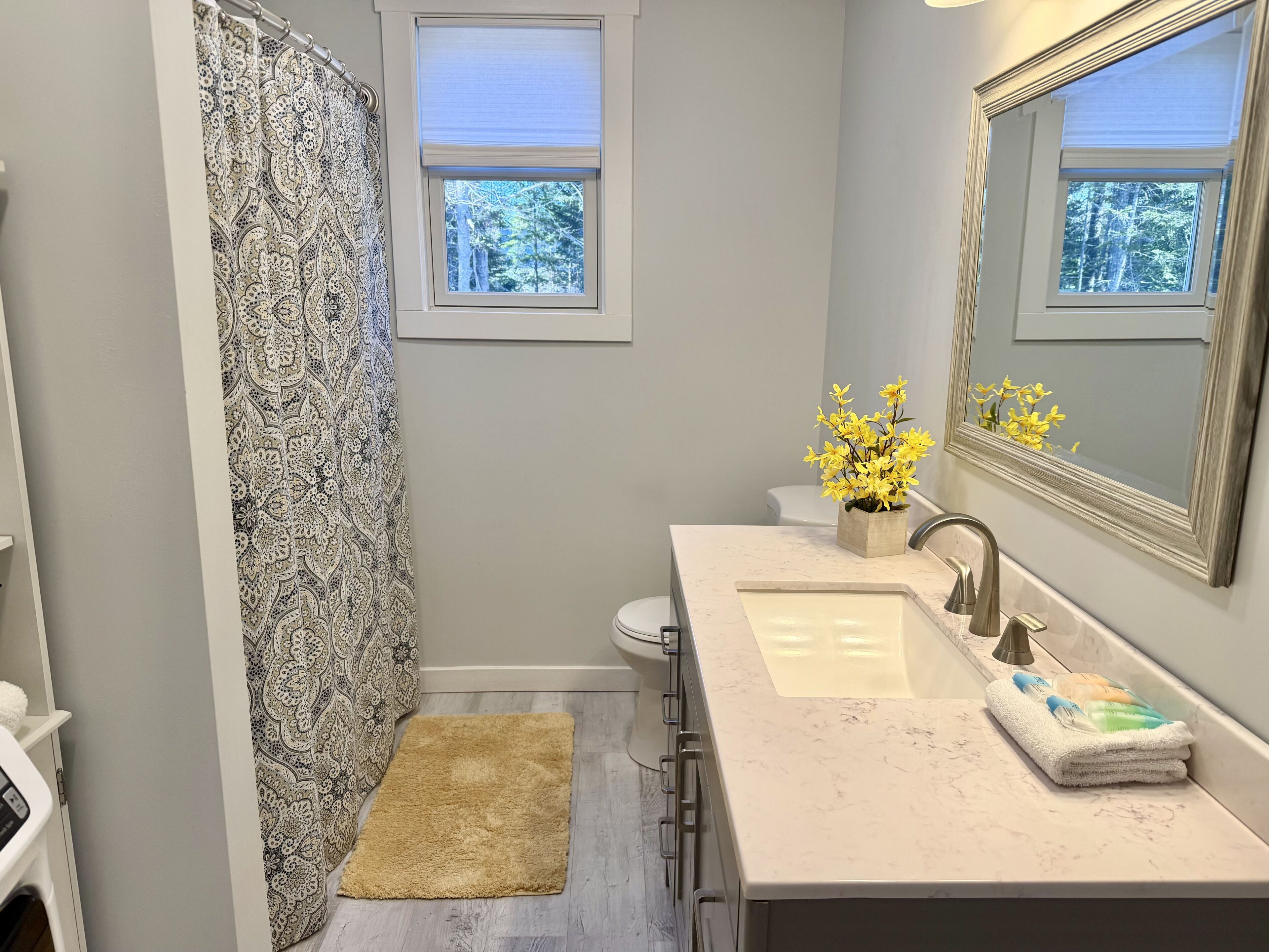 A bathroom with a shower curtain, a toilet, a sink with a mirror, and a window.