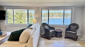 A room with a sofa, two wicker chairs, a TV, and large windows with a view of a lake and forest. - Waterfront Cottage on Private Island near Acadia (Milbridge)