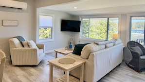 A living room with a sofa, armchair, TV, and large windows with a view of trees and a lake. - Waterfront Cottage on Private Island near Acadia (Milbridge)