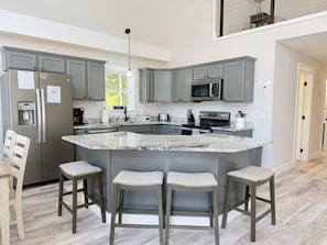 Uma cozinha moderna com armários cinza, uma ilha central com banquetas, uma geladeira de aço inoxidável e um micro-ondas.