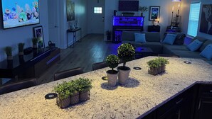 Un salón moderno con isla de cocina, lámparas colgantes azules y un televisor que muestra una escena de naturaleza.