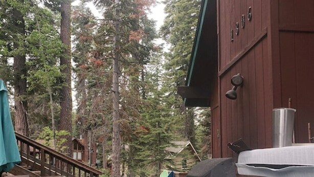 A hot tub in a forested area with a wooden deck and a large red building in the background. - Quiet & Large house - Hot Tub, Pool table, Fast WiFi  -  close to Kings Beach! (Tahoe Vista)