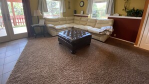 Una sala de estar con un sofá seccional color beige, un reposapiés marrón oscuro, una mesita de noche con lámpara y una ventana con cortinas.