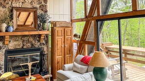 A cozy cabin interior with a stone fireplace, a large chandelier made of antlers, and a view of the forest through large windows. - FLAT, Lakefront chalet; Hottub, Playground, Firepit, Wifi, and more! (Wright City)