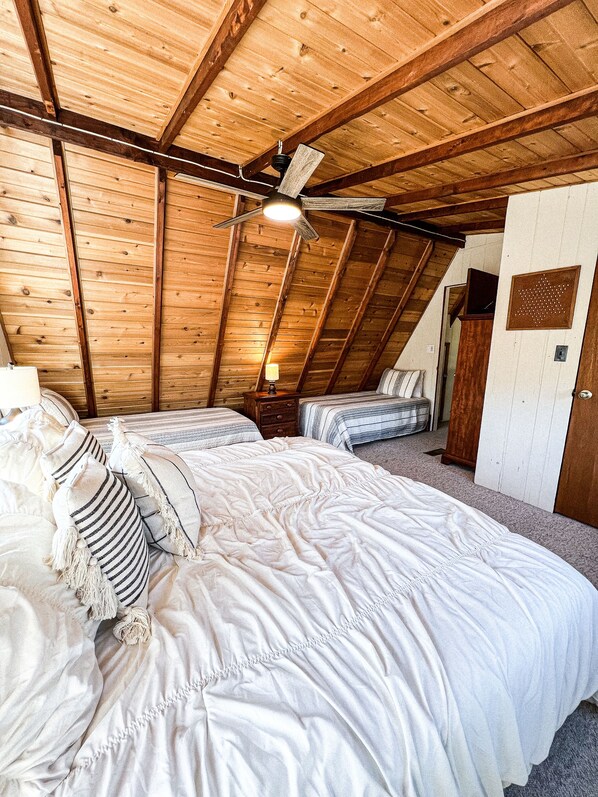 A bedroom with a sloped ceiling, wooden beams, a bed with white bedding, a nightstand, and a ceiling fan. - FLAT, Lakefront chalet; Hottub, Playground, Firepit, Wifi, and more! (Wright City)