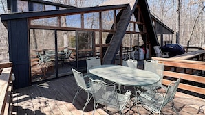 A wooden deck with a glass-enclosed dining area, metal chairs, and a table. - FLAT, Lakefront chalet; Hottub, Playground, Firepit, Wifi, and more! (Wright City)