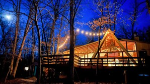 A wooden cabin with a triangular roof, surrounded by trees and string lights. - FLAT, Lakefront chalet; Hottub, Playground, Firepit, Wifi, and more! (Wright City)
