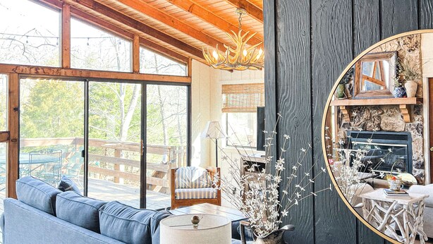 A cozy living room with a blue sofa, a wooden ceiling, and a large window overlooking a deck. - FLAT, Lakefront chalet; Hottub, Playground, Firepit, Wifi, and more! (Wright City)