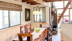 A dining area with a wooden table and chairs, a hanging light fixture, and a view of the outdoors through large windows. - FLAT, Lakefront chalet; Hottub, Playground, Firepit, Wifi, and more! (Wright City)