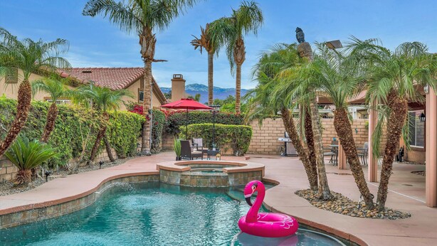 A swimming pool with a pink flamingo float, surrounded by palm trees and a patio area with outdoor furniture. - Spacious Indio Oasis - Pool & Spa near Polo Ground (Indio)