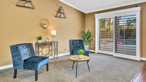 A room with two blue upholstered chairs, a round coffee table with a plant, and a large window with a view of a pool and palm trees. - Spacious Indio Oasis - Pool & Spa near Polo Ground (Indio)