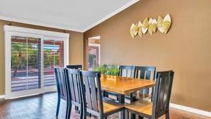 A dining area with a wooden table and black chairs, a brown wall with decorative leaf art, and a sliding glass door leading to an outdoor area with plants. - Spacious Indio Oasis - Pool & Spa near Polo Ground (Indio)