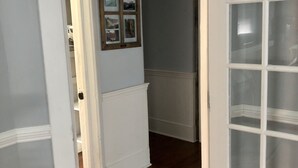 A hallway with a wooden floor, a white door with glass panels, and a wall with framed pictures. - The Apollo (Charleston)